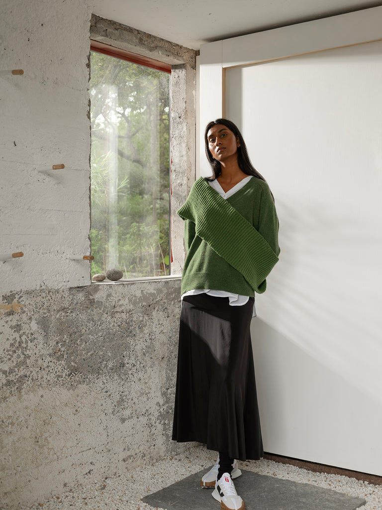 Full length portrait of dark skinned and long dark haired woman wearing green v neck sweater with white shirt underneath, green knit scarf, black skirt and white trainers standing beside window in white room