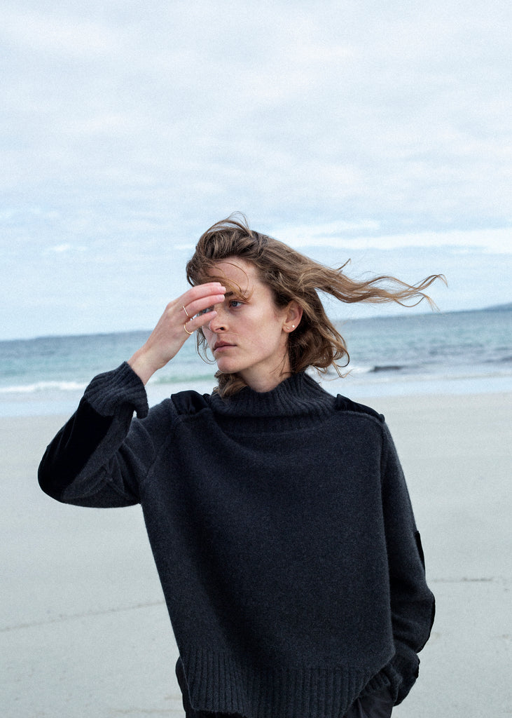  Upper body photo of white female on West of Ireland shoreline wearing Irish designed luxury cashmere sweater in a Charcoal mélange colour from Loro Piana. It has shoulder epaulettes and long patches on the lower sleeves made from Black velvet.