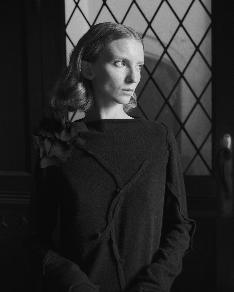 Black and white portrait of a woman wearing a back cashmere sweater with rose corsage in a room with geometric window design.