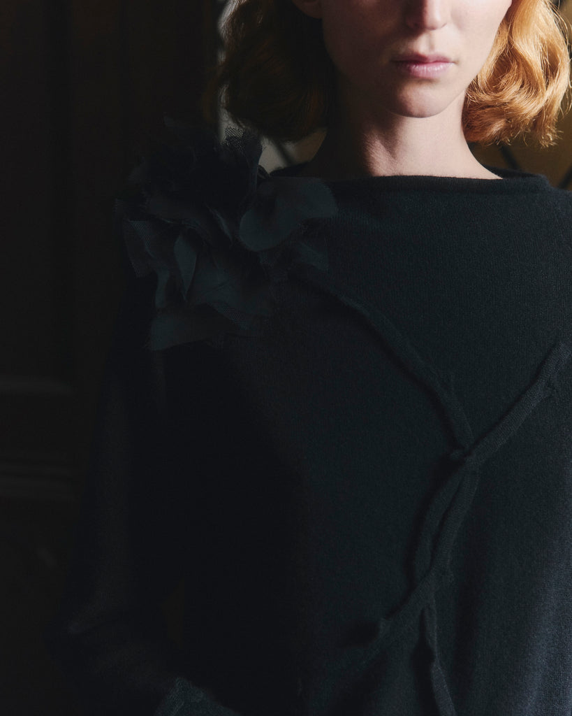 Woman with red hair wearing a black cashmere swearer with rose corsage against a blurred background