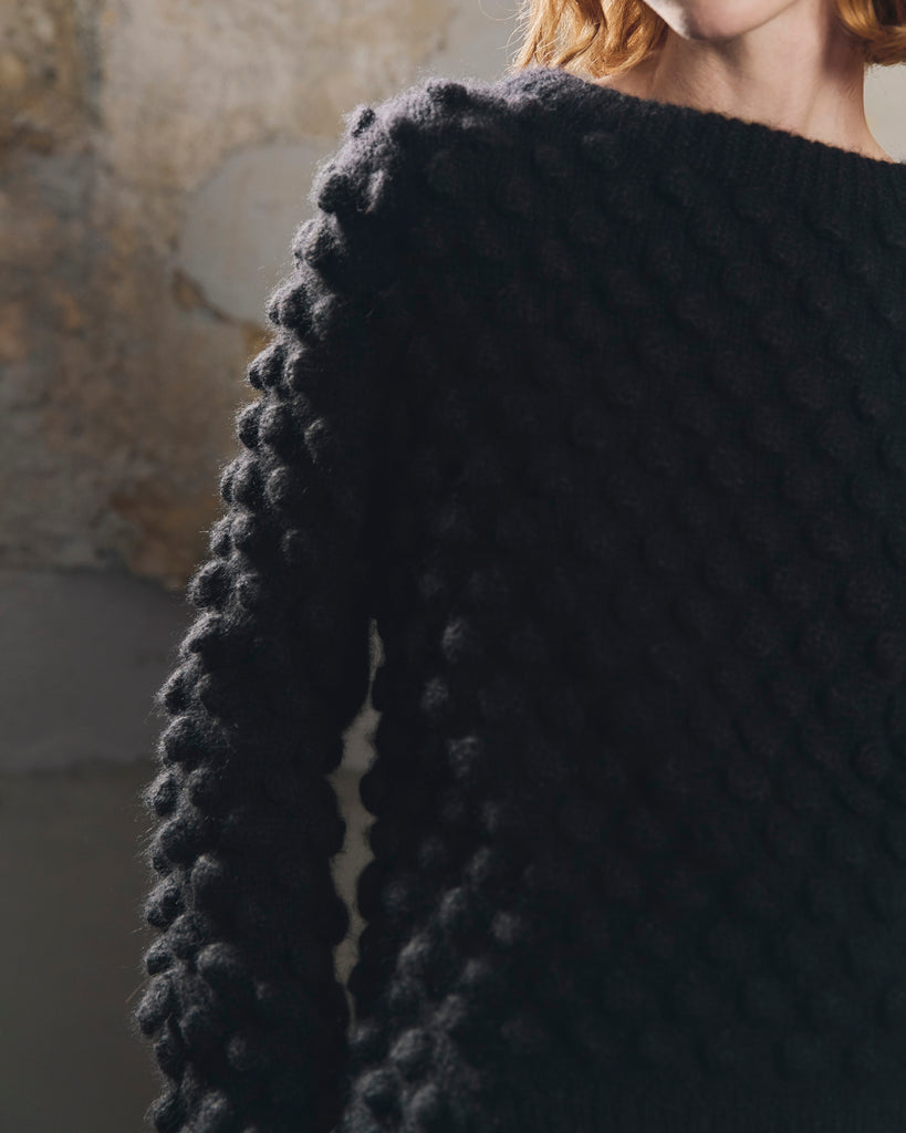 Close-up of a woman with red hair wearing a black chunky knitted sweater with a textured bobble pattern.