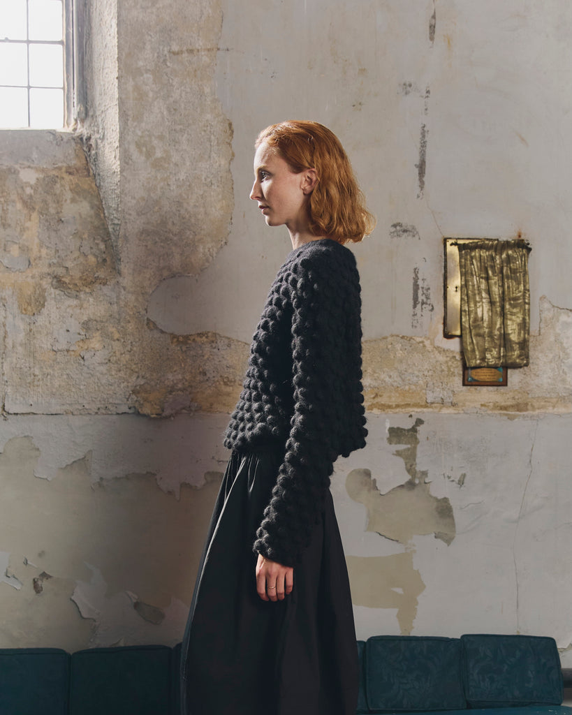 Woman with red hair wearing a black hand knit bobble pattern textured sweater and skirt against a wall with peeling paintwork and high window