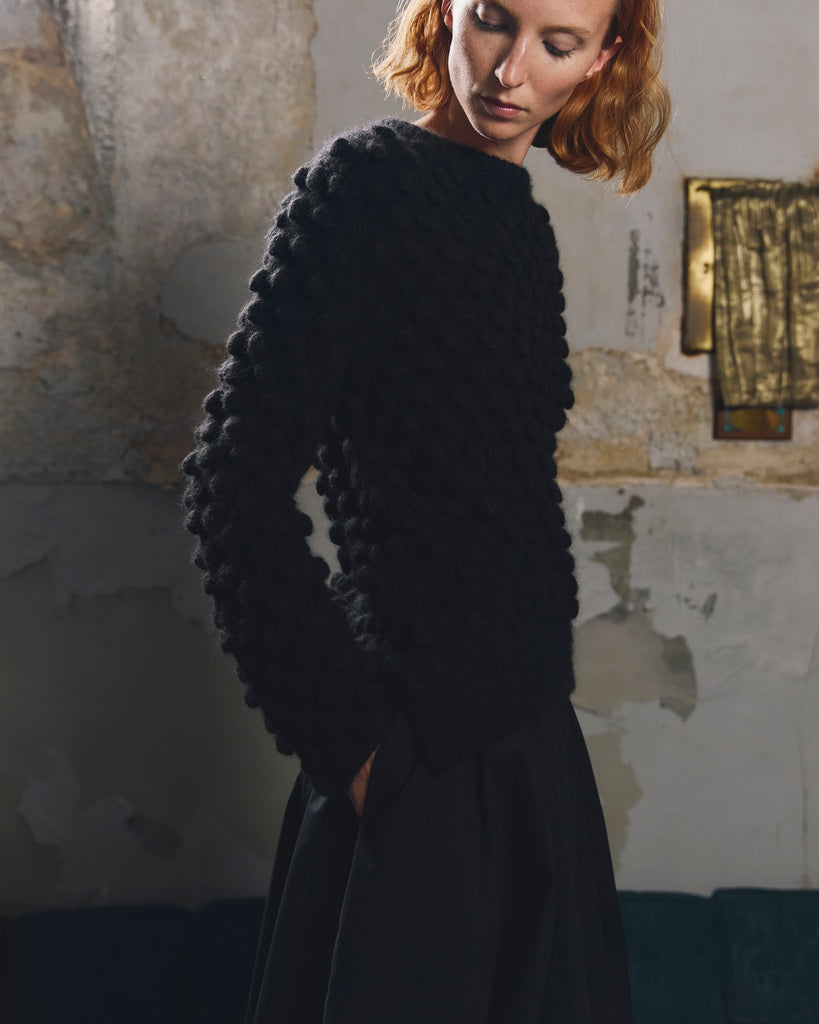 Woman with red hair and hands in pocket of a black skirt wearing a black textured baby alpaca hand knitted bobble sweater against a wall with peeling paintwork and decorative gold detail