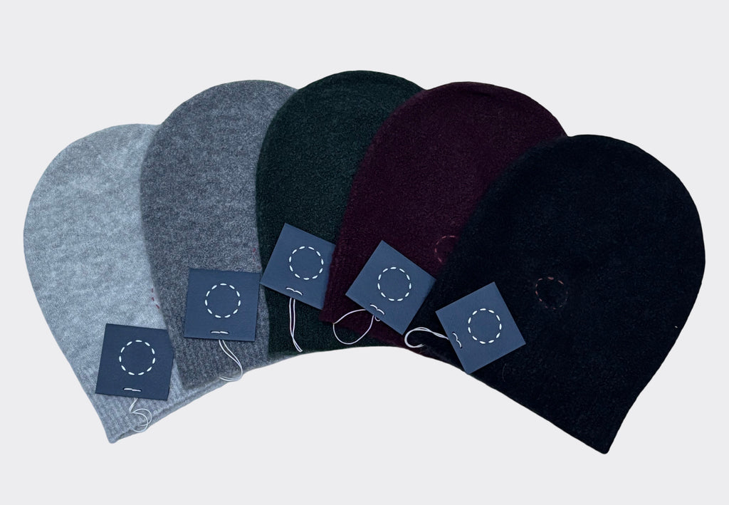 Product image of five cashmere silk beanie hats in gray, light grey, black, burgundy and first green on a light gray background