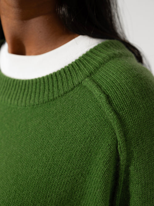 Cropped photo of woman with dark hair and brown skin wearing green round neck Sphere One cashmere sweater with white top underneath only the side of neck and chin is visible