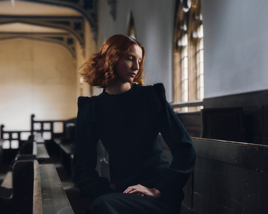 Woman with red hair wearing black cashmere sweater sitting in church pew with large window in background