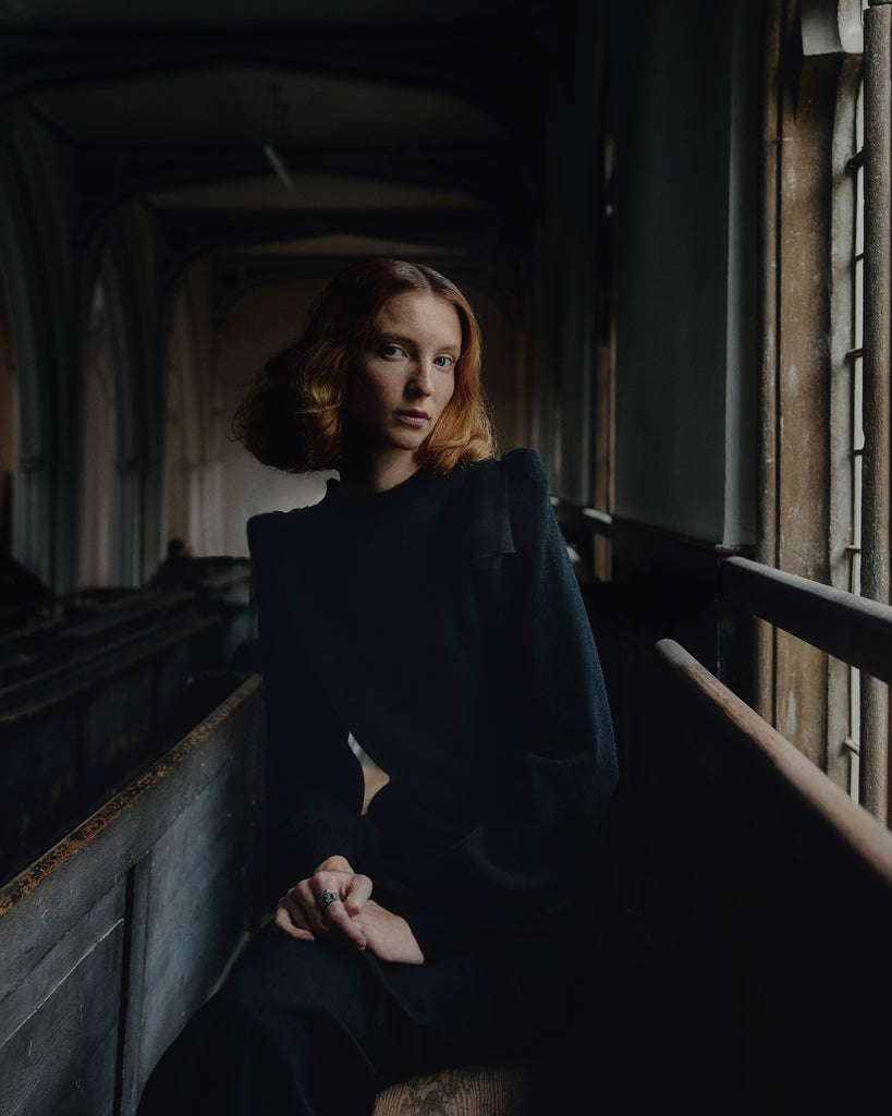 Woman with red hair wearing black cashmere sweater sitting with hands clasped in a dimly lit room with a light falling on her face from large window