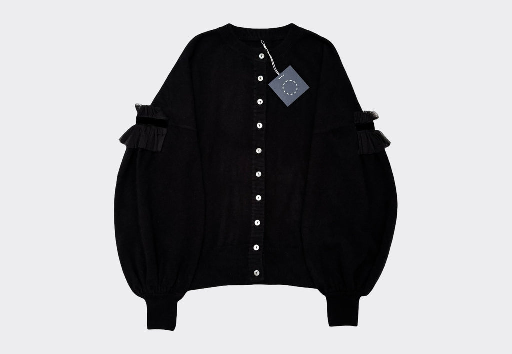 Black round neck cardigan with light coloured buttons, dropped shoulder and bulbous lower sleeves with tulle velvet detail on a light gray background