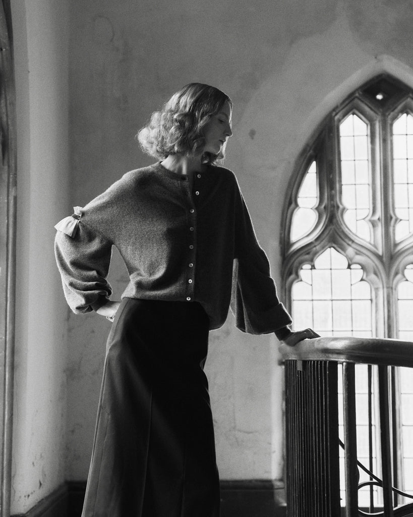 Black and white photo of a woman with her hand on her hip wearing a cashmere cardigan standing in a room with gothic architecture