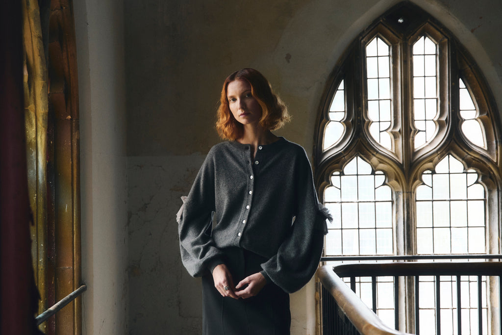 Red haired woman wearing a grey dropped shoulder cashmere cardigan with bulbous sleeves standing with hands clasped  in a room with large gothic windows