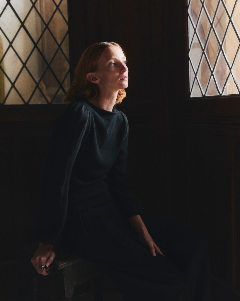 Woman with red hair wearing black cashmere and chiffon Sphere One sweater sitting in a dimly lit room with large windows