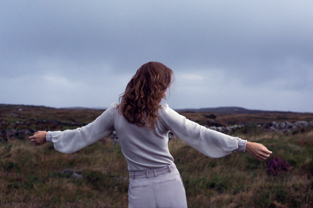 Person standing in a field with heather and stone walls with arms outstretched, looking towards a cloudy sky.