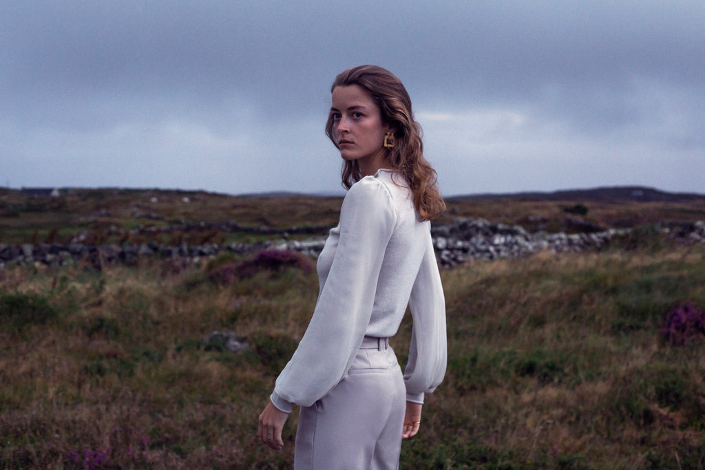 Woman in a grey silver cashmere outfit standing in a grassy heather and stone wall field with a cloudy sky.