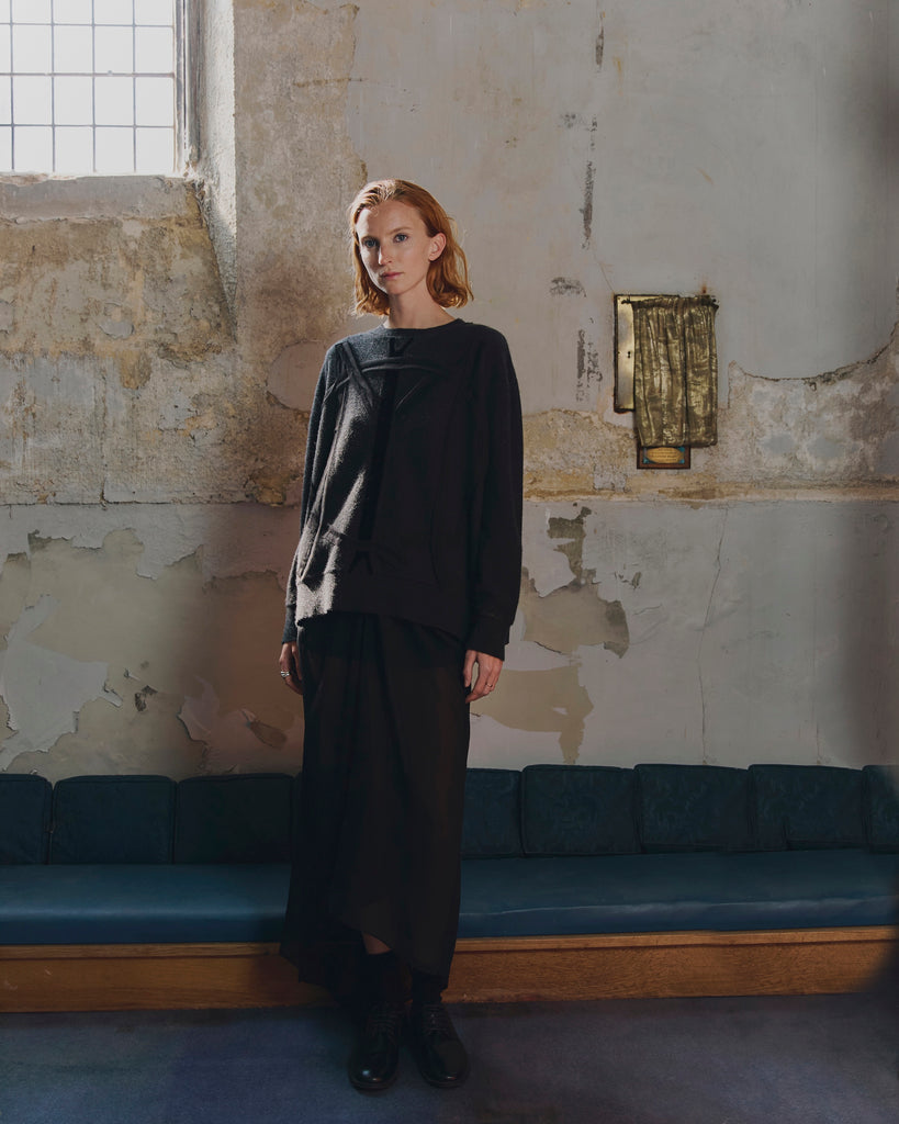 Woman with red hair in a dark grey cashmere sweater and black outfit standing in a room with peeling paint and a blue cushioned bench.