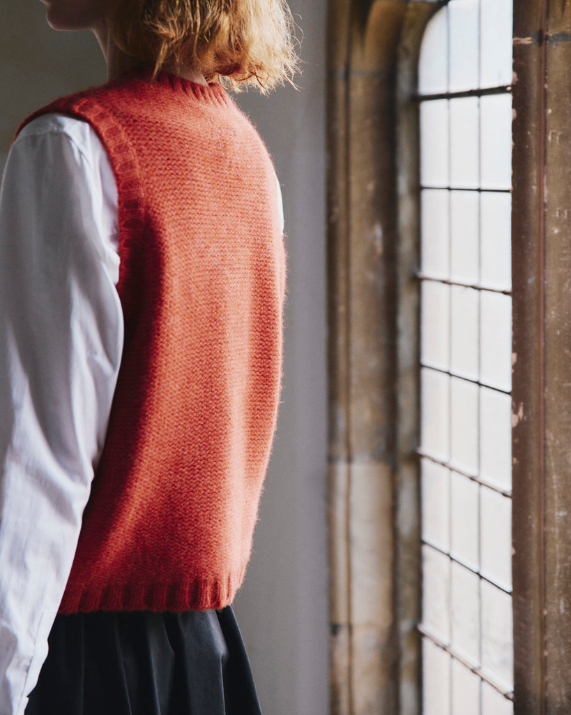 Person with red hair wearing a red knitted vest over a white shirt in a room with large windows.