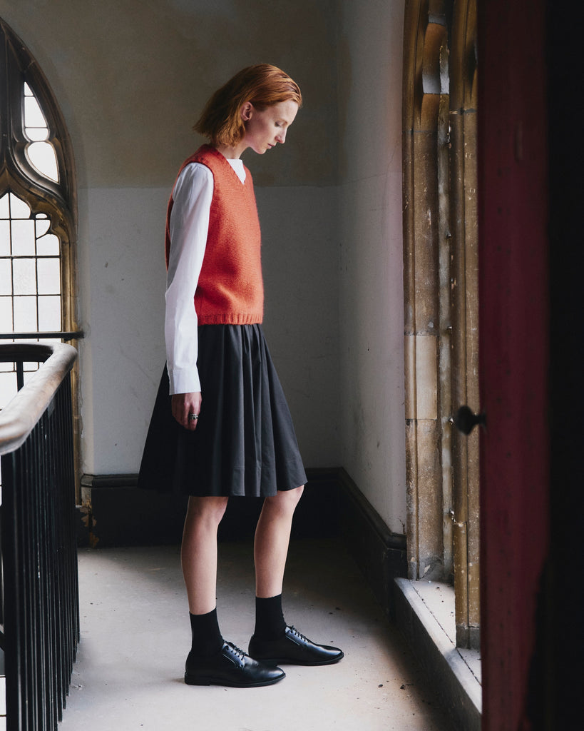 Woman with red hair wearing a red v neck knitted sweater vest over a white shirt and black skirt facing a large ornate gothic window in a dimly lit room.