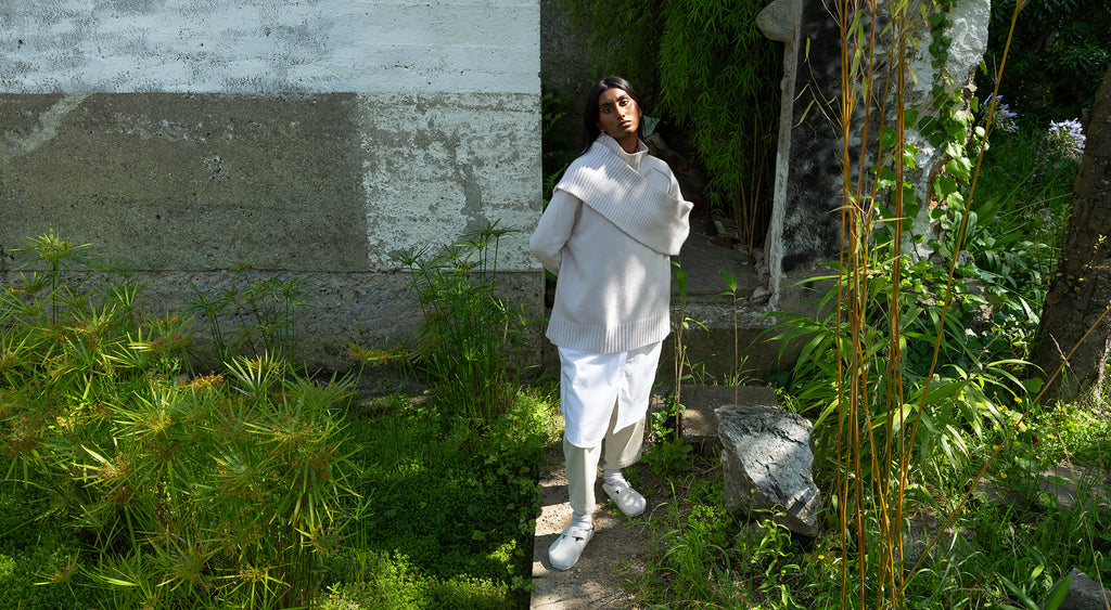 Woman wearing white knitted Sphere One cashmere sweater, scarf across her torso, with dark skin and black hair, standing with her arms behind her back in front of white painted architectural concrete structure surrounded by green plants