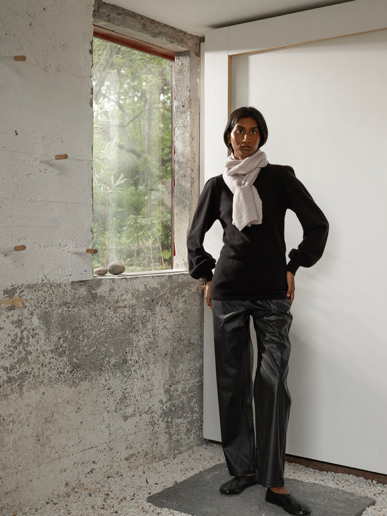 Full length image of woman with dark hair and skin looking directly at camera standing beside window with hands on her hips wearing black cashmere knit sweater and white scarf and black leather effect trousers