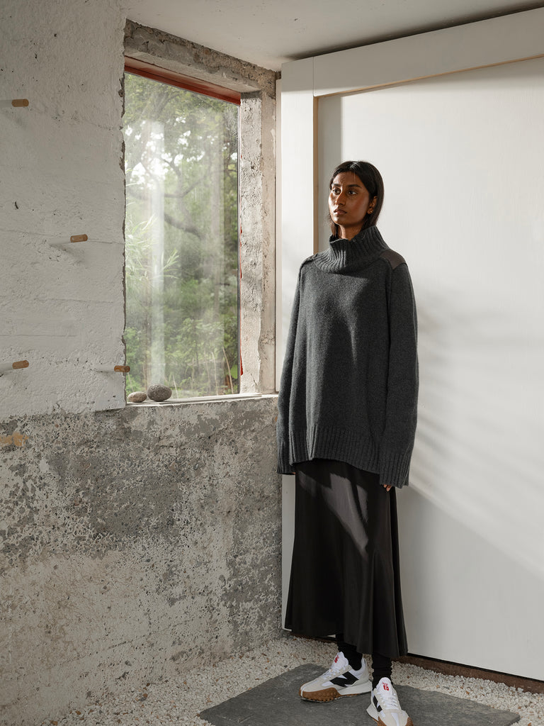 Woman with dark hair standing beside window in a white and grey room wearing oversized high neck cashmere sweater with black long skirt and white trainers