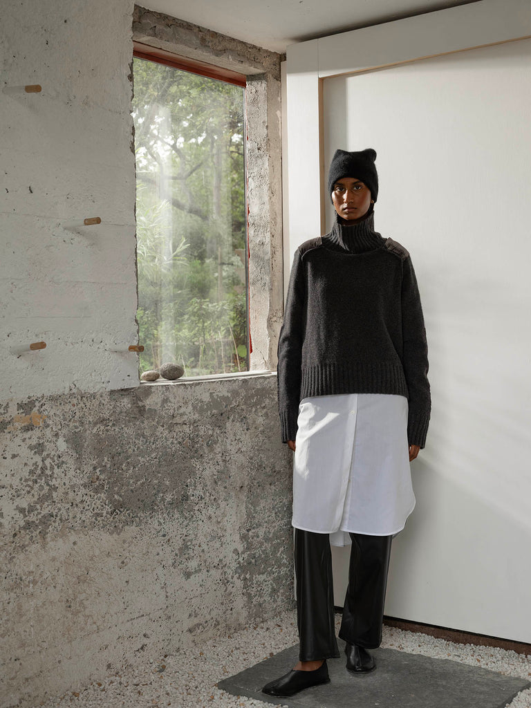 Full length portrait of dark skinned woman looking at camera wearing high neck earthy green colour cashmere sweater with hat, long white shirt, black trousers standing beside window in white room
