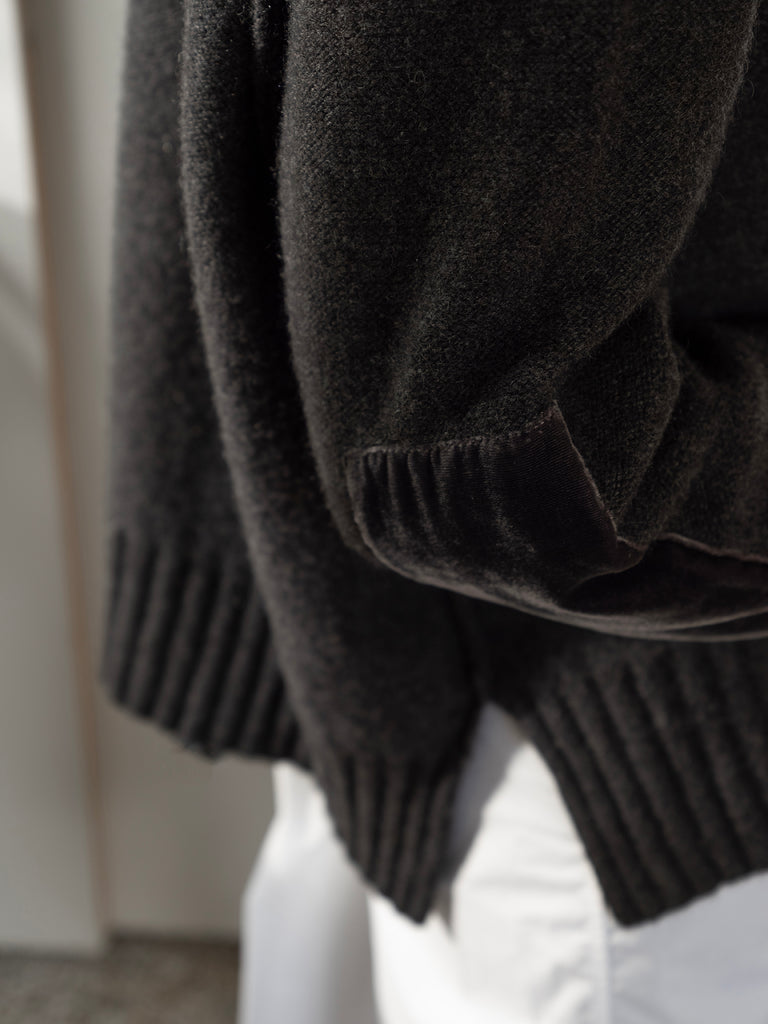 Close up image from side of model wearing earthy green cashmere sweater with velvet elbow patch detail with rib split hem worn with white trousers