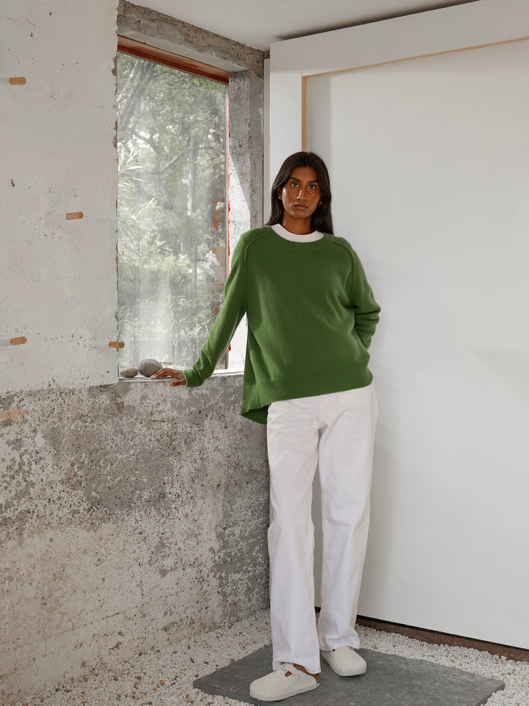 Dark skinned model wearing green cashmere round neck sweater with white top underneath and white trousers standing with one hand on windowsill in a white room