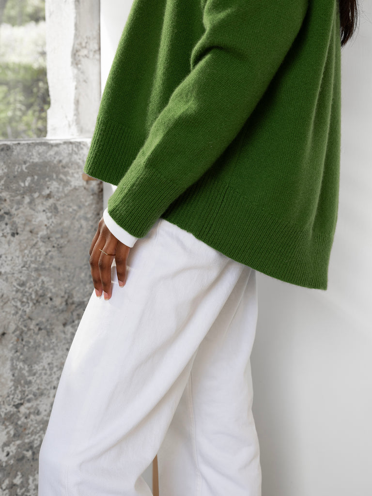 Side view close up of dark haired female model wearing green cashmere sweater with curved hem and white trousers, hand placed on her thigh, standing beside window