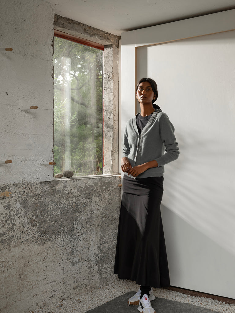 Dark skinned model wearing grey cashmere cardigan, long black skirt and white trainers standing beside window in a white and concrete room