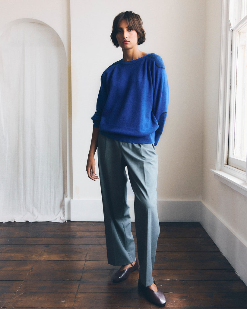 A model stands beside a window in a white room with a wooden floor, one arm behind her back and one foot forward she is looking directly at the camera. She has short brown hair and is wearing a blue round neck cashmere sweater with patches on the arms and elbows. One sleeve is pushed up. She is also wearing grey pants and burgundy Mary Jane style flat shoes. 