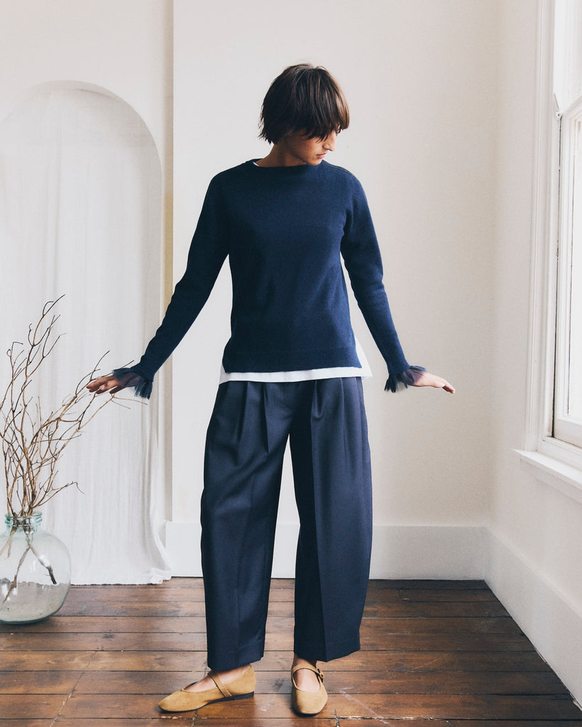 A woman stands looking at a window in a white room with a wooden floor. She has short brown hair and is wearing a navy cashmere sweater with navy chiffon cuffs and navy trousers. Her ands are splayed out at her sides. There is a glass vase on the floor with some branches displayed in front of a white curtain in an arched doorway.