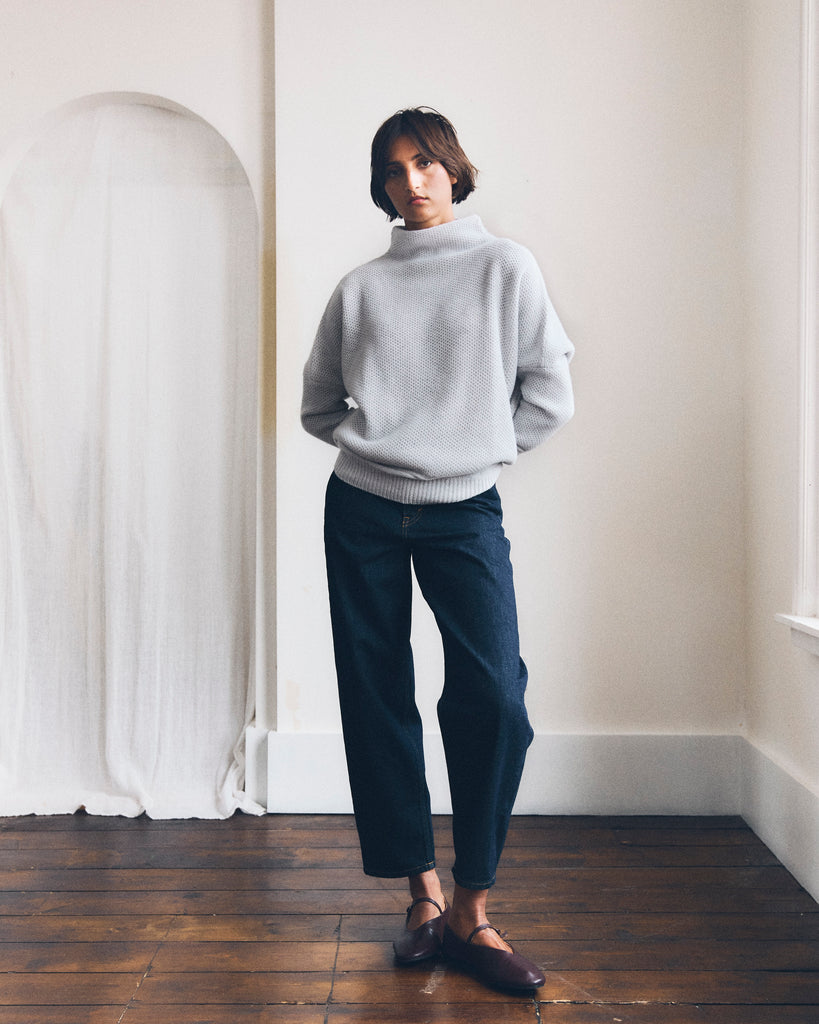 A model stands beside a window in a white room with wooden floors and a curtained archway, her hands behind her back she looks at the camera. She has short brown hair and is wearing a grey honeycomb texture cashmere sweater from Sphere One and dark jeans cropped at the ankle. 