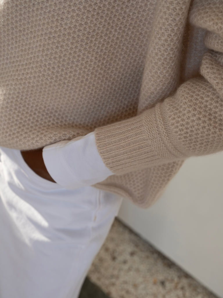 Detail lookbook shot of model with brown skin wearing honeycomb pattern cream cashmere sweater with rib cuff with hand in pocket of white trousers Irish knitwear brand Sphere One