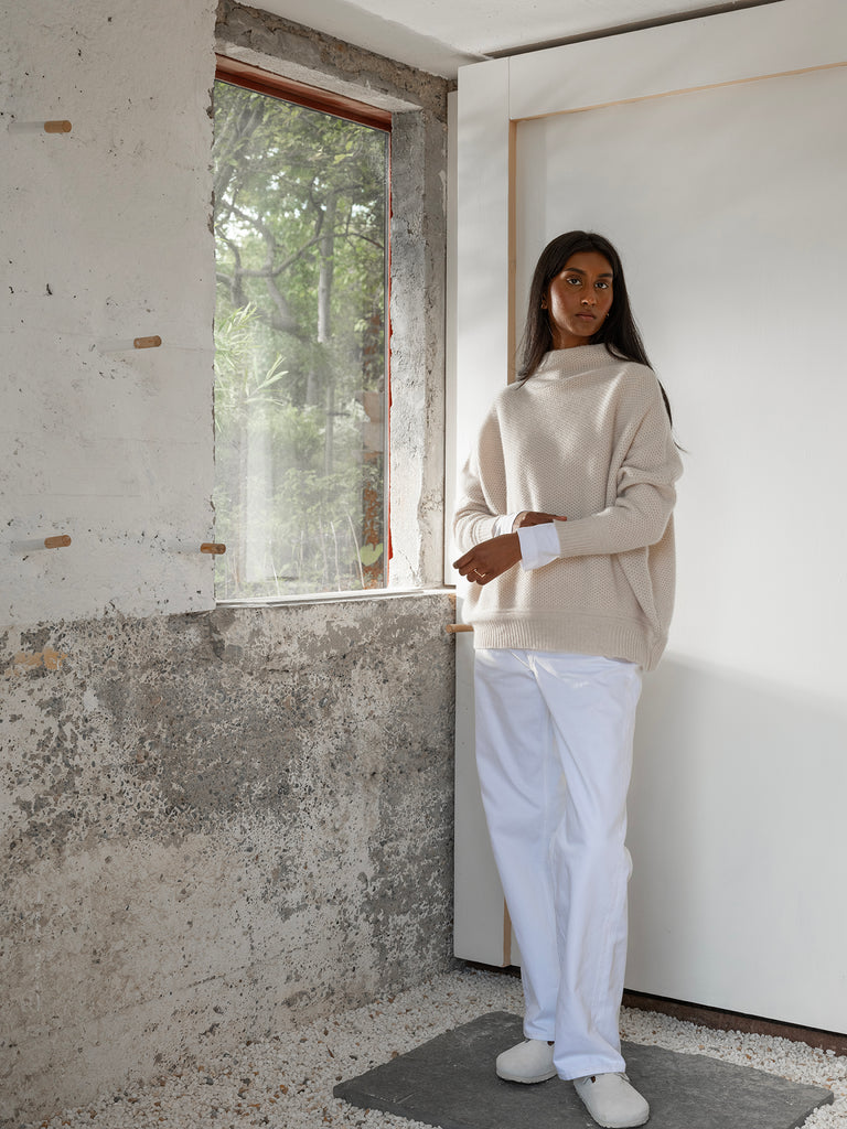 Portrait lookbook shot of model standing beside a window against white and grey concrete room with brown skin and long black hairwearing honeycomb pattern cream cashmere sweater with rib cuff with white trousers and white shoes Irish knitwear brand Sphere One