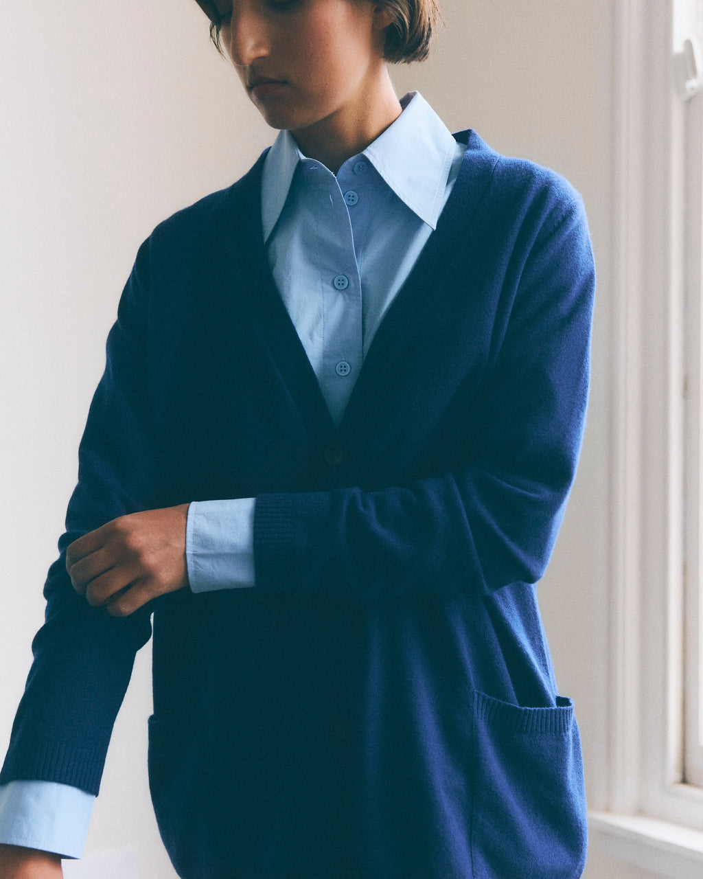 Woman with short dark hair models a blue v neck cashmere cardigan, she is also wearing a light blue shirt with wide lapels 