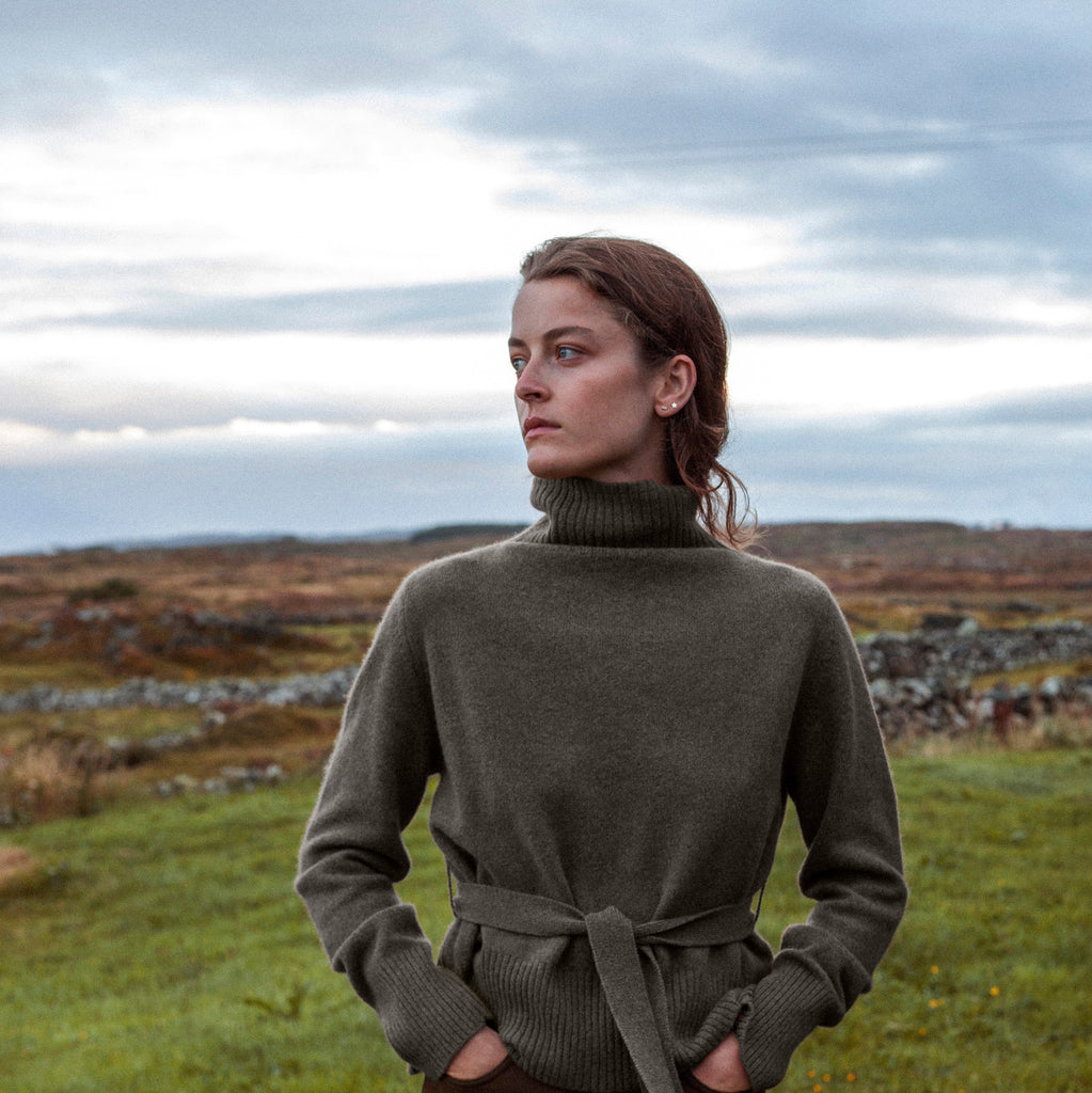 Brown haired woman looking to the left with hands in pockets wearing belted high neck cashmere sweater against Irish landscape and sky